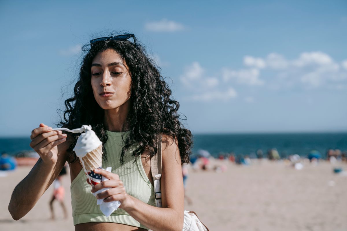 Mujer en la playa comiendo un helado de cucurucho con una cuchara, con el mar y la arena de fondo, justo antes de notar el efecto de comer helado rápido.
