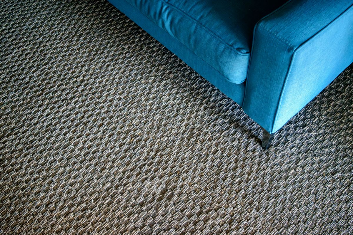 Pata metálica de un sofá azul apoyada sobre una alfombra tejida de fibra natural, dejando una marca de muebles en la trama.