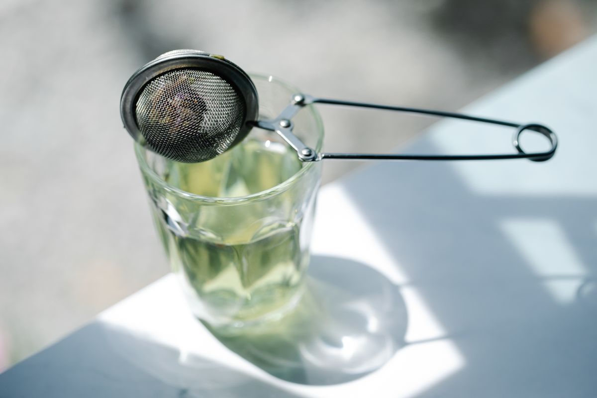 Colador de té de metal enganchado al borde de un vaso de cristal con líquido dentro, bajo la luz del sol.