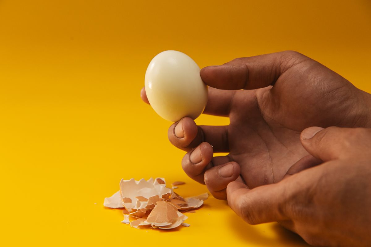 Mano sosteniendo un huevo duro ya pelado junto a los trozos grandes de cáscara, resultado del truco del bicarbonato para pelar huevos duros, sobre fondo amarillo.