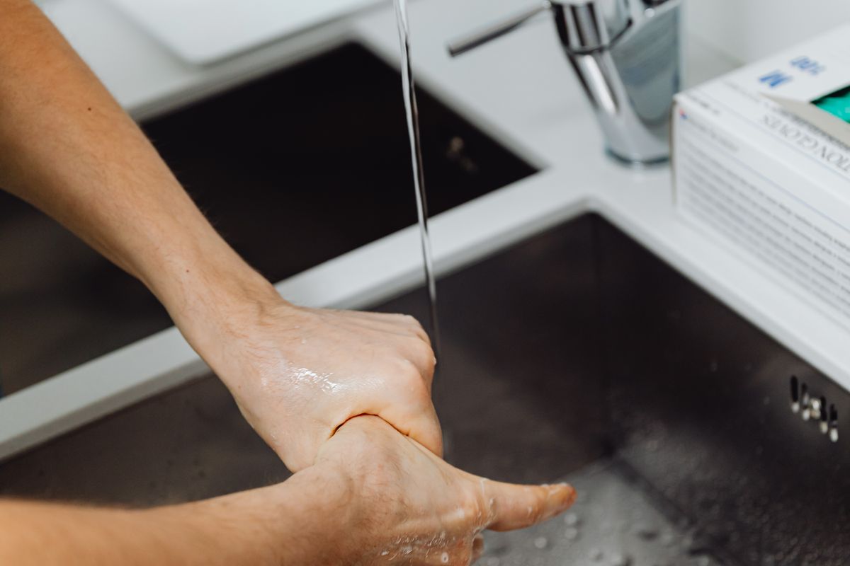 Unas manos se frotan bajo el agua fría del grifo de acero inoxidable de una cocina, con una caja de guantes cerca del fregadero.