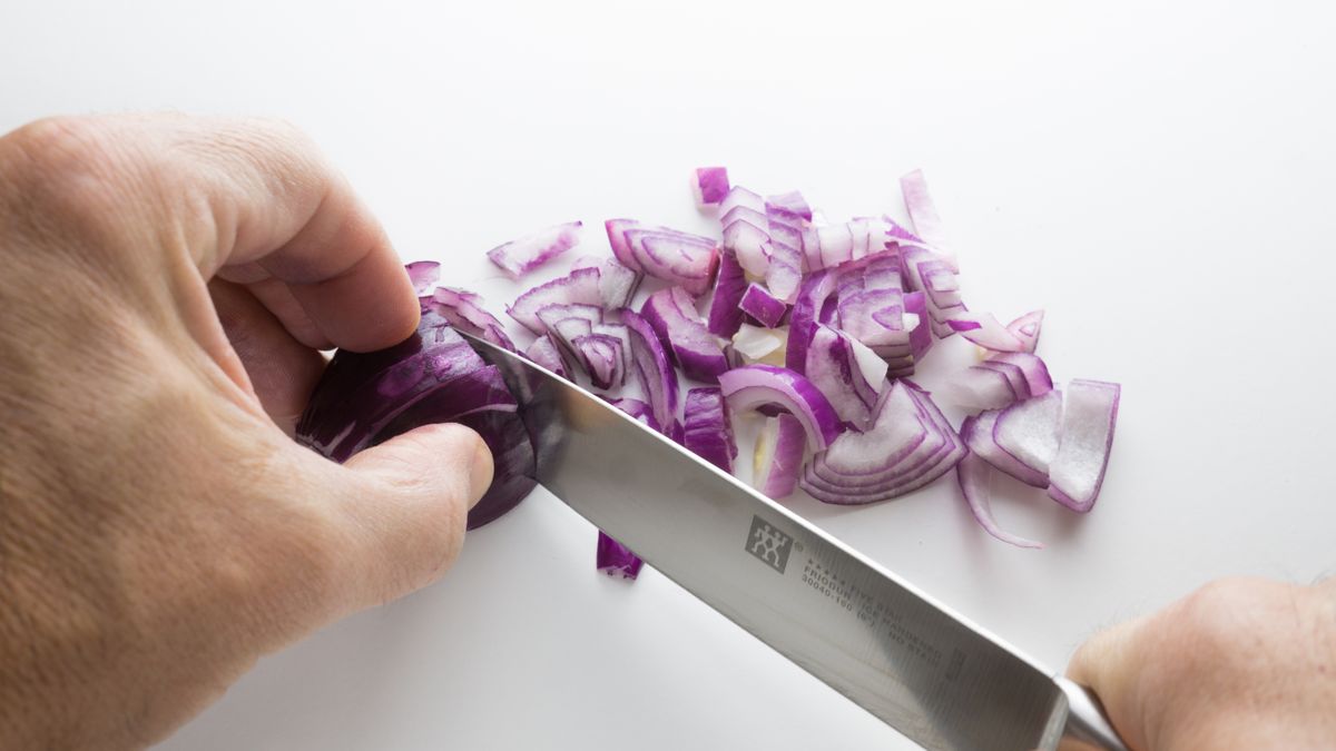 Manos cortando una cebolla morada en trozos con un cuchillo de cocina sobre una tabla blanca.