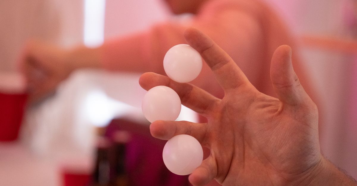 Una mano sostiene tres pelotas de ping-pong blancas, con vasos rojos de fiesta desenfocados al fondo.