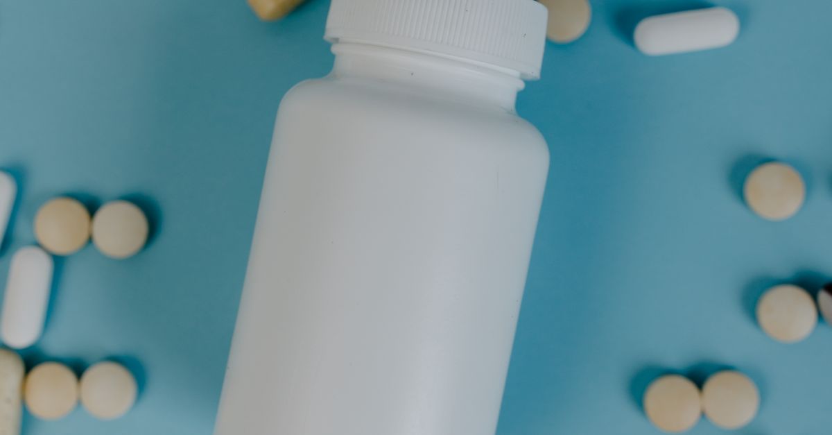 Bote blanco de pastillas junto a comprimidos beige y cápsulas blancas esparcidos sobre un fondo azul.