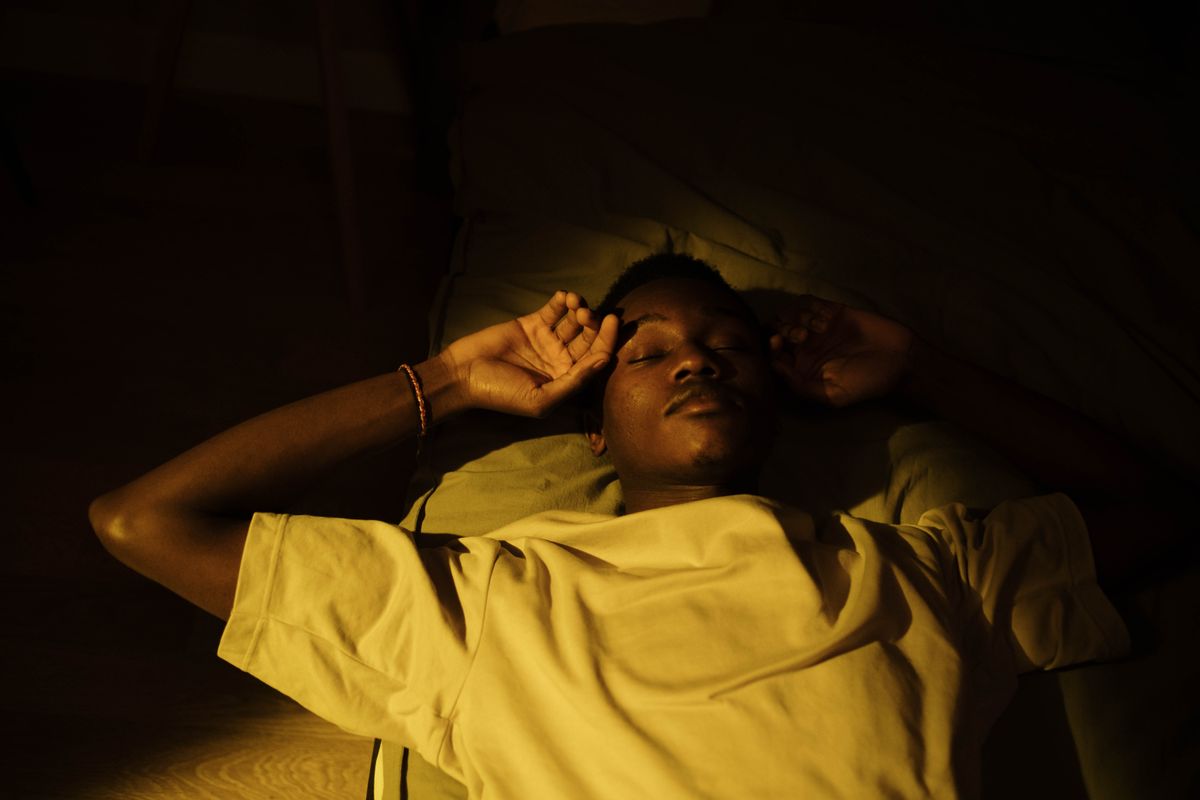Hombre joven tumbado en la cama con los ojos cerrados y una mano cerca de la frente, iluminado por una luz cálida y tenue, en el instante de despertar y empezar a olvidar los sueños.