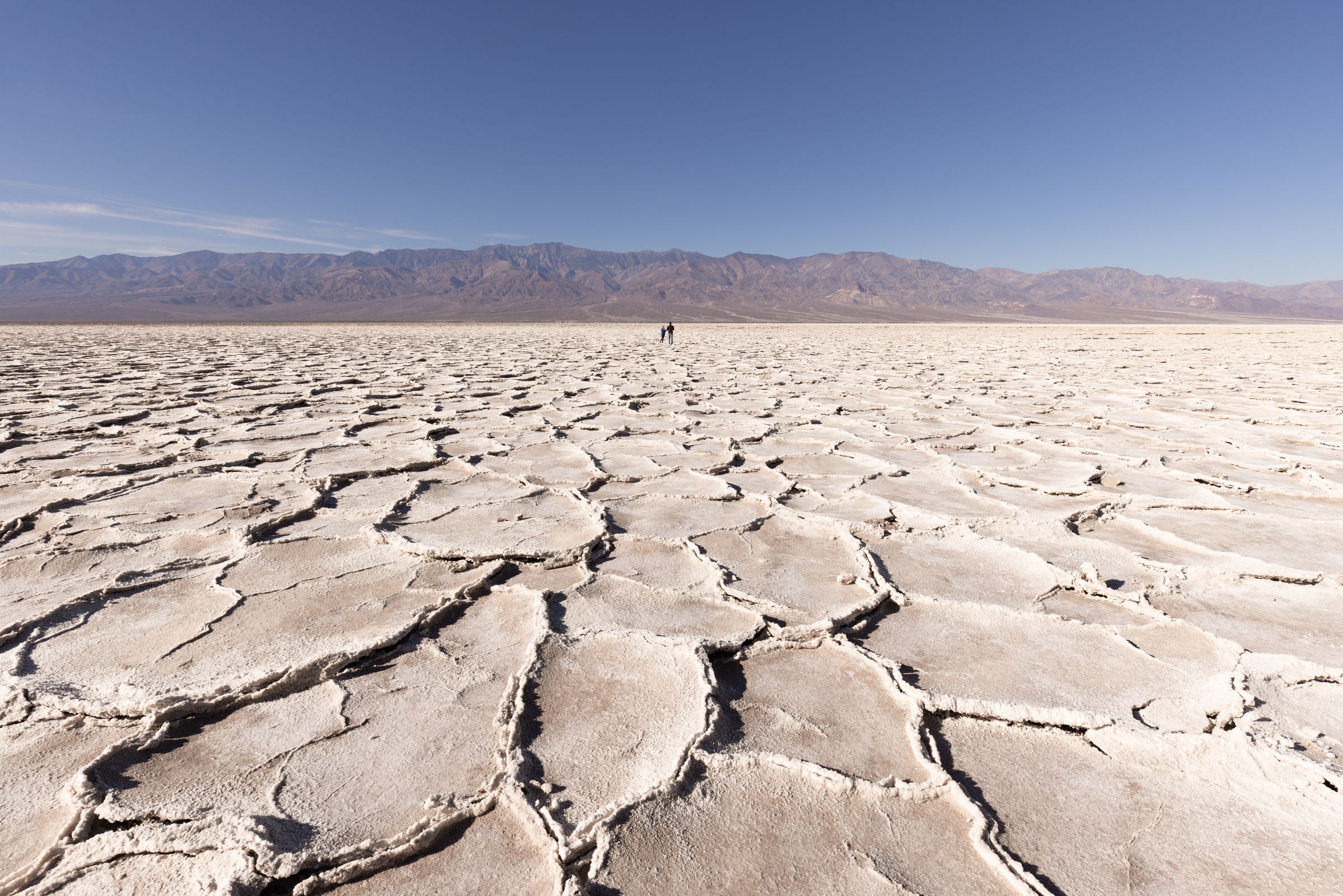 Vista panorámica del suelo agrietado y blanco de un lago seco en Death Valley, con montañas al fondo y dos personas caminando en la distancia.
