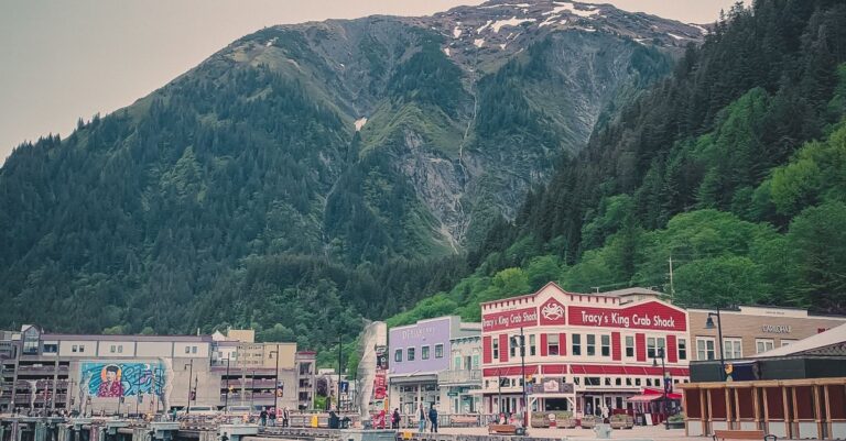 Pueblo costero de Alaska con casas de colores y el letrero de un restaurante de cangrejos, rodeado de montañas boscosas y un fiordo.