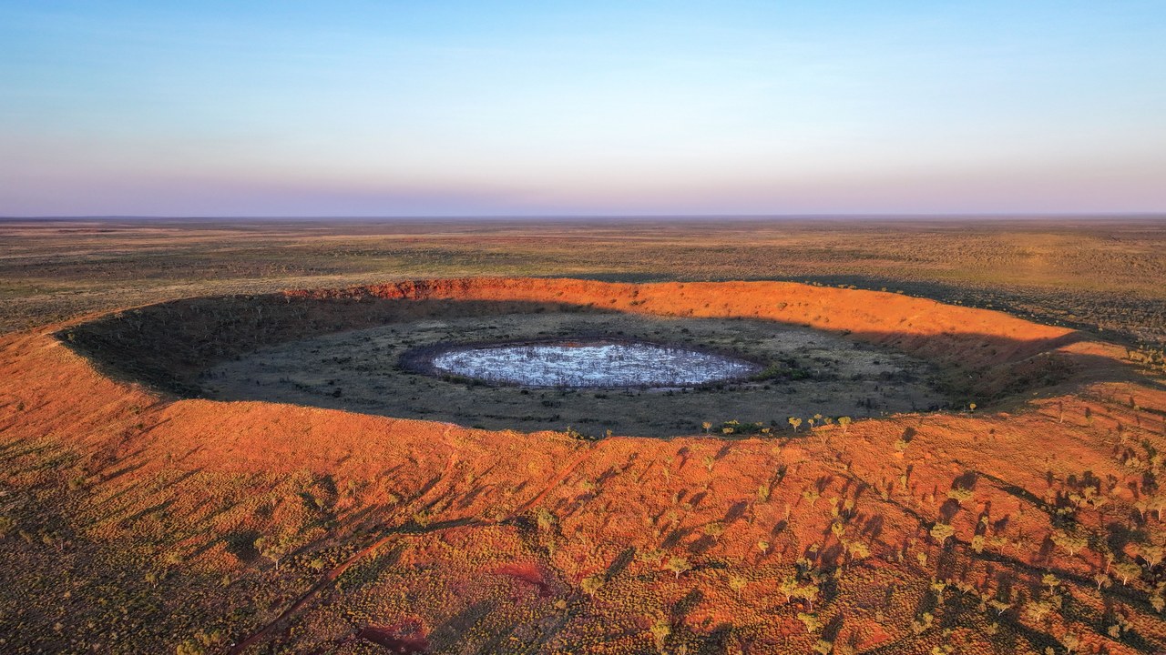 Vista aérea de un cráter de meteorito circular, con paredes de tierra rojiza y un suelo claro en el centro, rodeado de llanura semiárida.